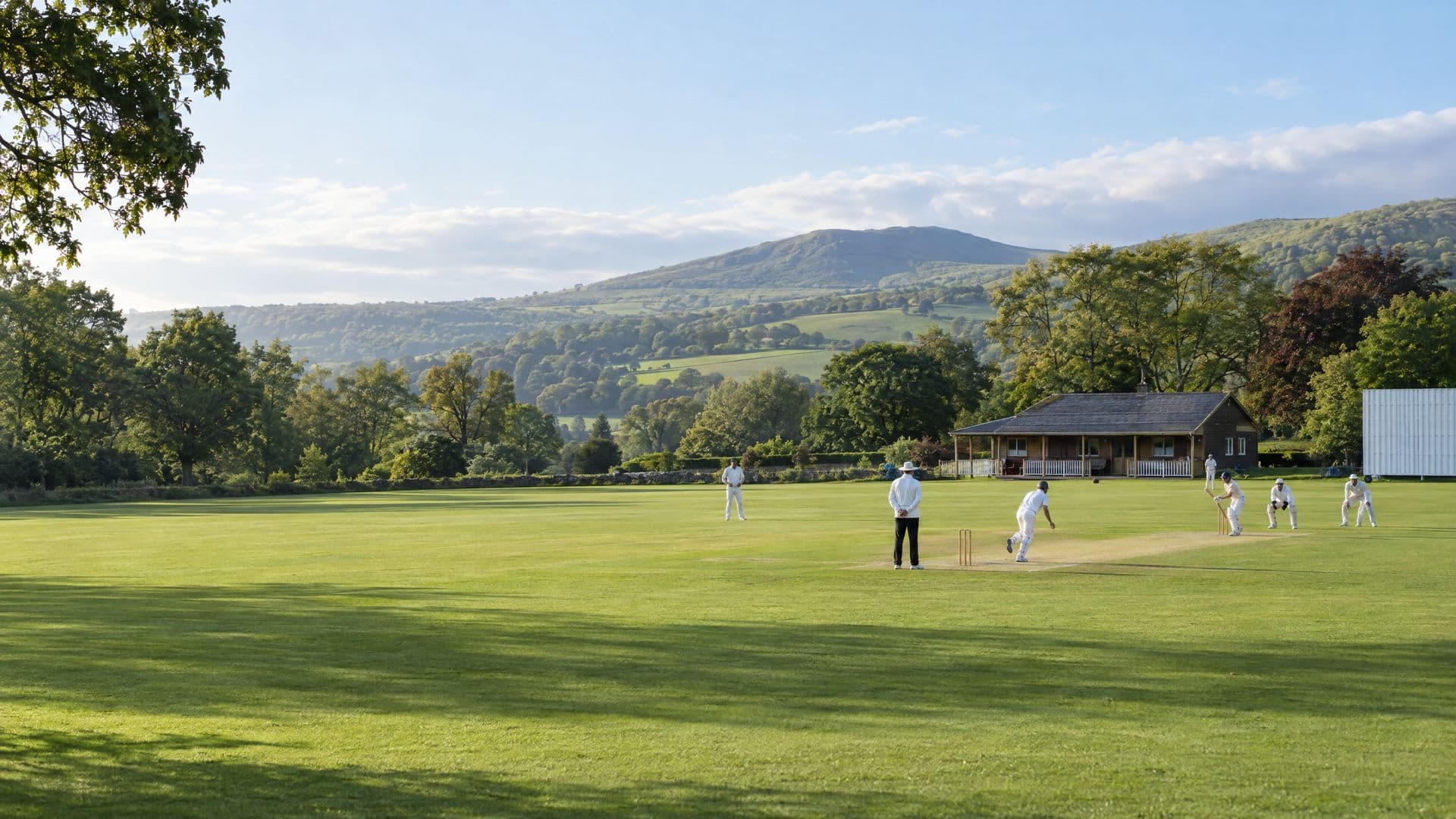 A cricket match on a green village ground, with a bowler running in, a wooden pavilion and wooded hills behind